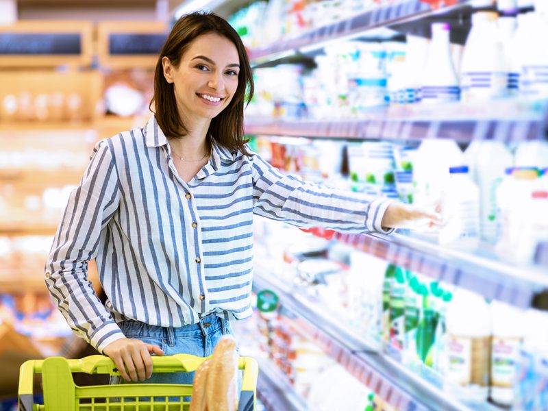 Retail Concept. Smiling Young Lady Standing With Shopping Trolley Cart Near The Shelves In Grocery Store. Cheerful Woman Buying Essentials In Super Market, Taking Dairy Products Looking At Camera
